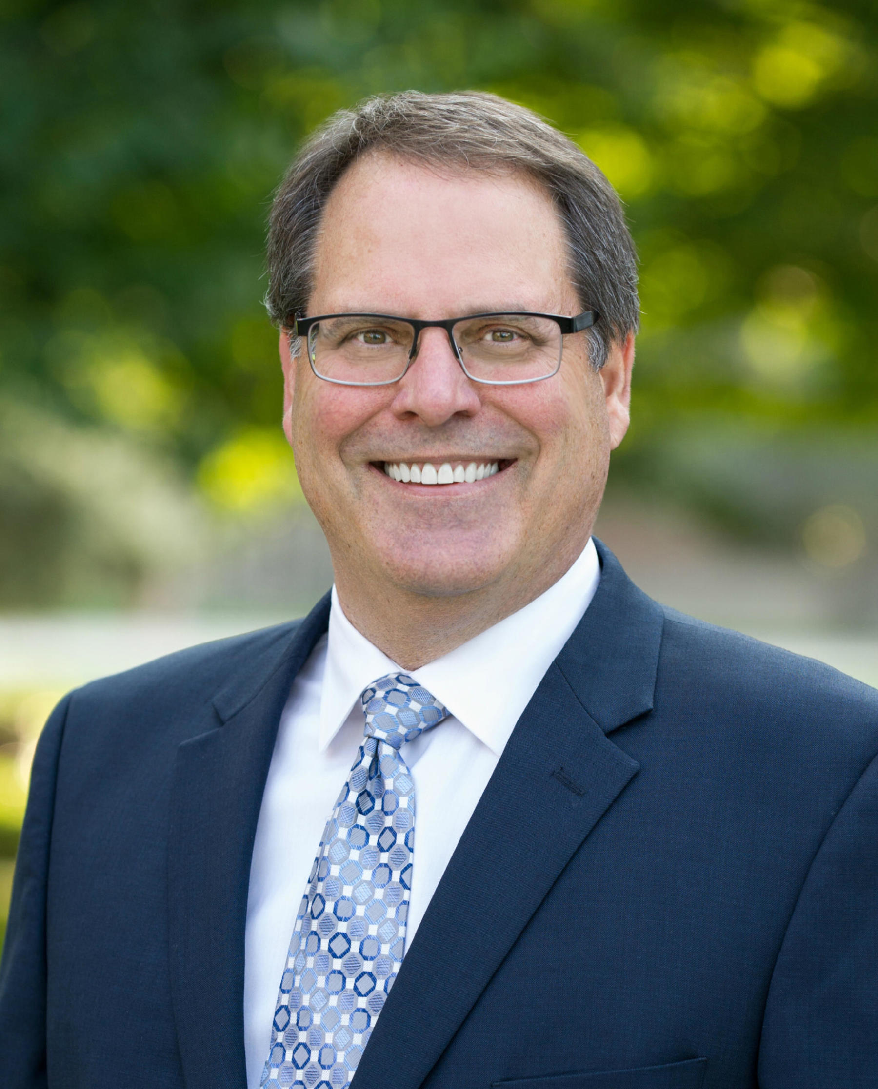 man outdoors smiling while wearing glasses and a suit coat and tie