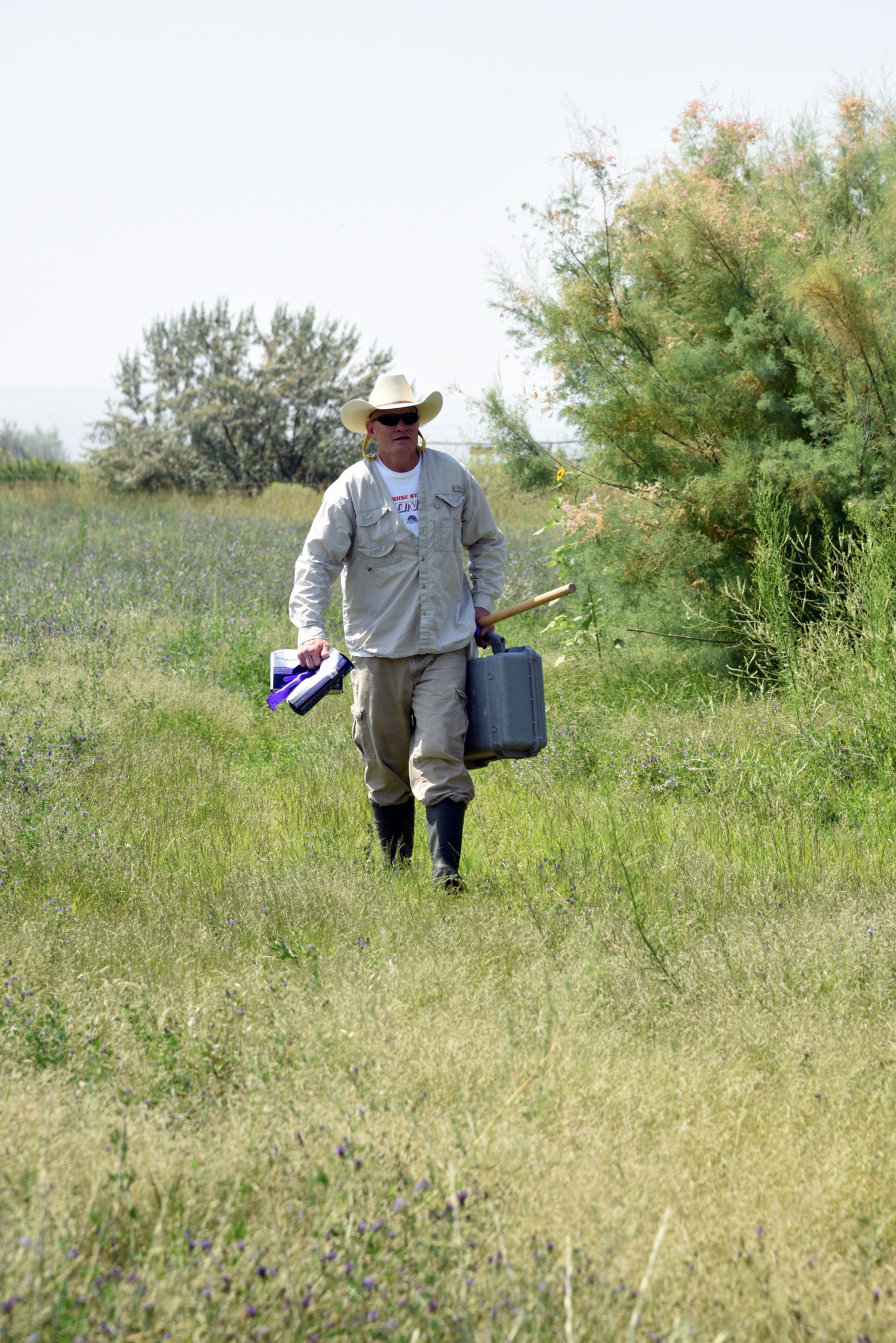 man walking through field wearing a cowboy hat carrying a plastic box